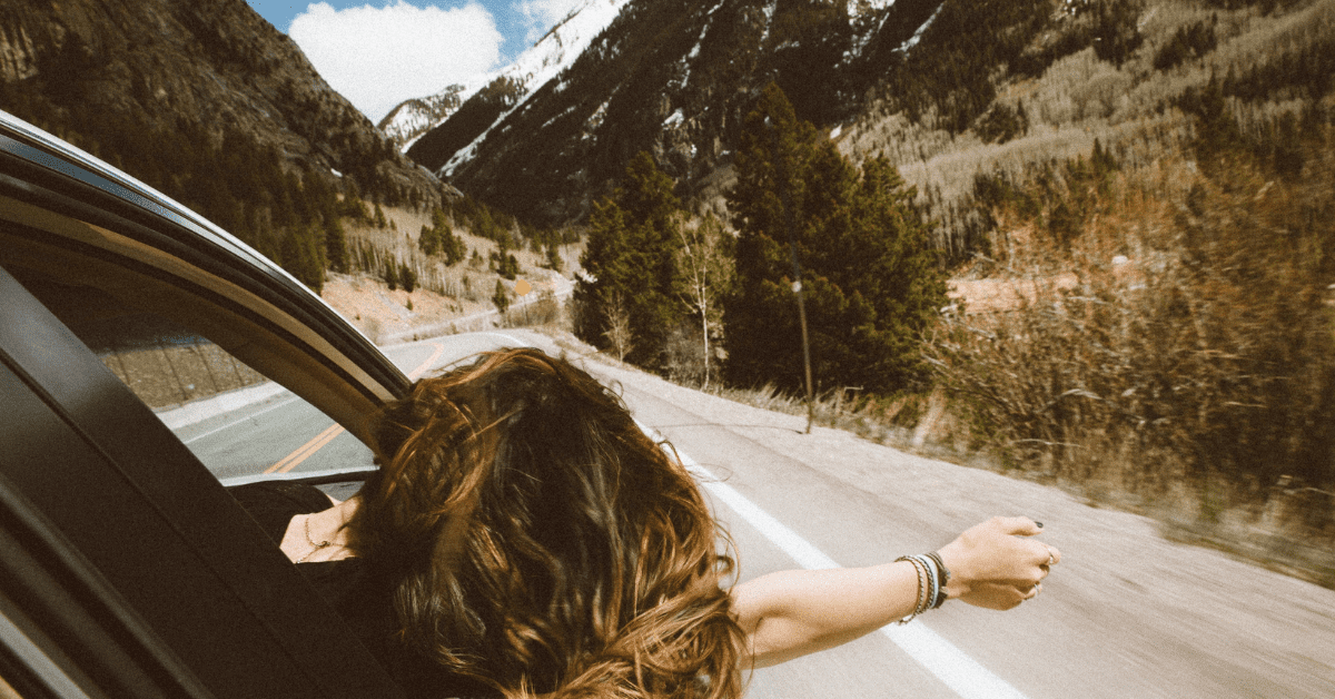 A woman reaches her arm out of the passenger window on a drive through the mountains.