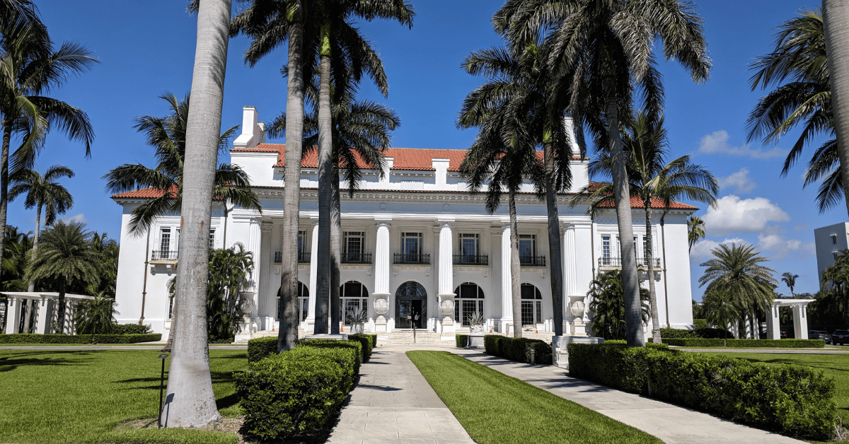 a white building with columns and a walkway with palm trees with Flagler Museum in the background