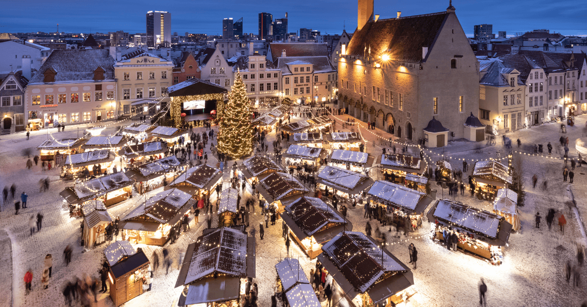 A festive Christmas market in a historic town square with snow-covered stalls, people, and a large tree.