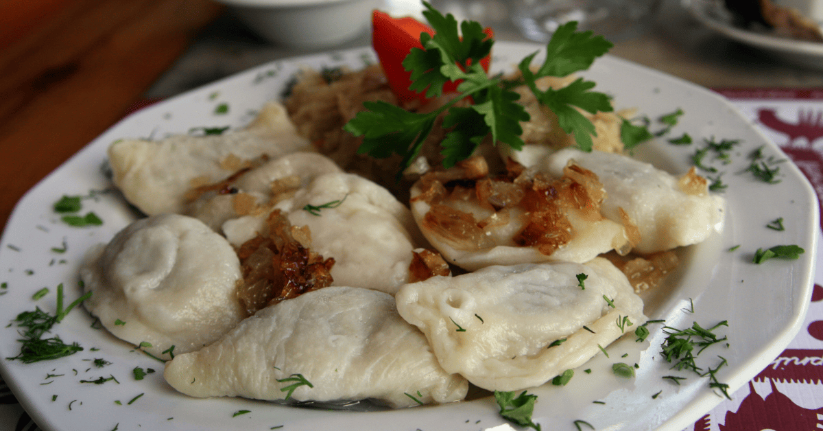 A plate of pierogi topped with caramelized onions, a sprig of parsley, and a slice of tomato, served on a white plate.