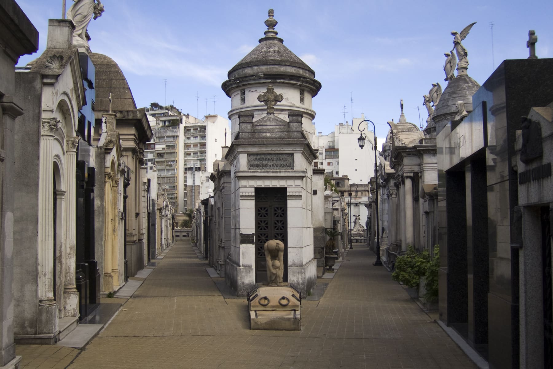 A serene and historical cemetery with ornate mausoleums, statues, and architectural details, set against a backdrop of modern buildings under a clear sky.