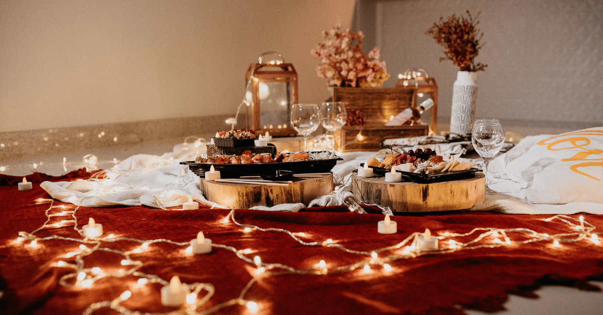 A cozy indoor picnic setup with assorted snacks on wooden platters, surrounded by fairy lights and candles, with wine glasses and decorative items enhancing the romantic ambiance.