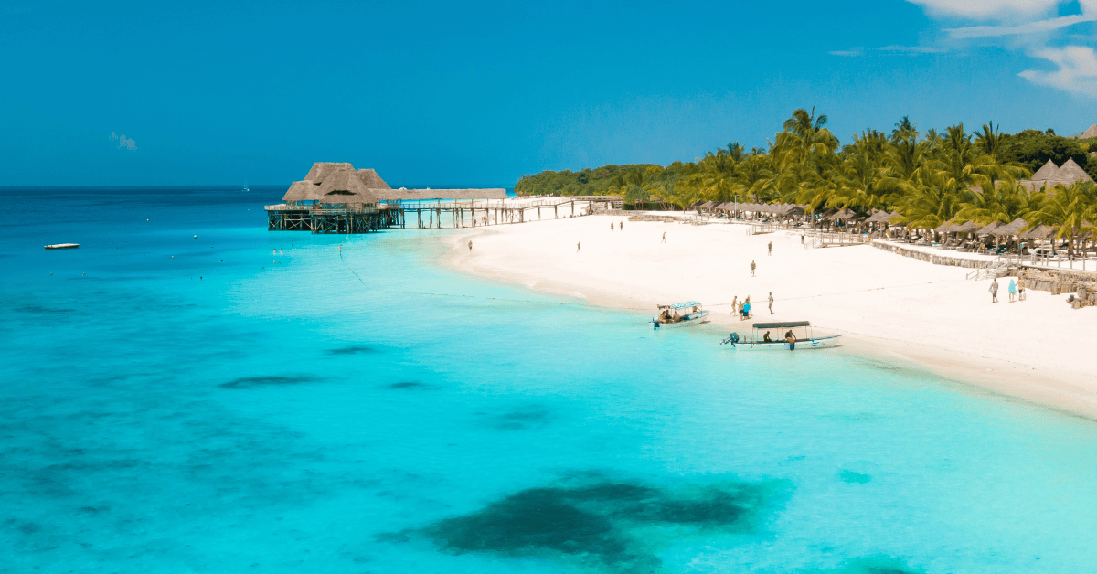 A serene beach with crystal clear turquoise waters, a white sandy shore, and a thatched-roof structure extending over the water under a clear blue sky.