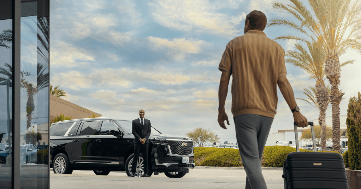 A casually dressed person with a grey suitcase is being greeted by a formally dressed chauffeur next to a black luxury car. Palm trees and a clear sky form the backdrop.