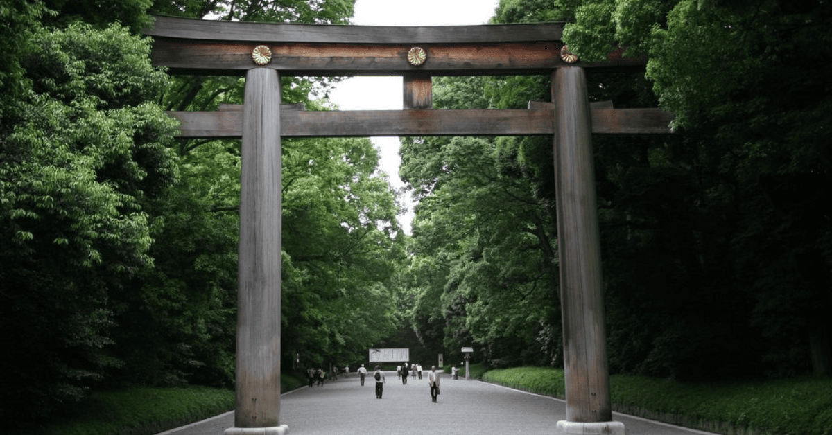 A majestic traditional wooden torii gate stands at the entrance of a serene path, flanked by lush green trees. A few people leisurely walk along the path, creating a tranquil scene. The gate’s intricate design and golden emblems evoke a sense of wonder and reverence. The natural canopy formed by the trees adds to the peaceful ambiance.