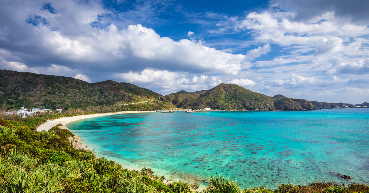 A breathtaking coastal landscape: turquoise sea, white sandy beach, and lush green mountains under a partly cloudy sky.