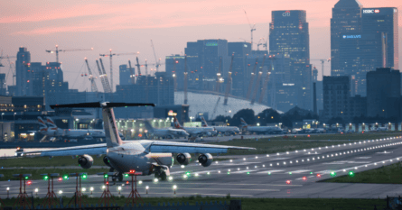 An airplane on a runway with the city skyline, illuminated by the setting sun, in the background.