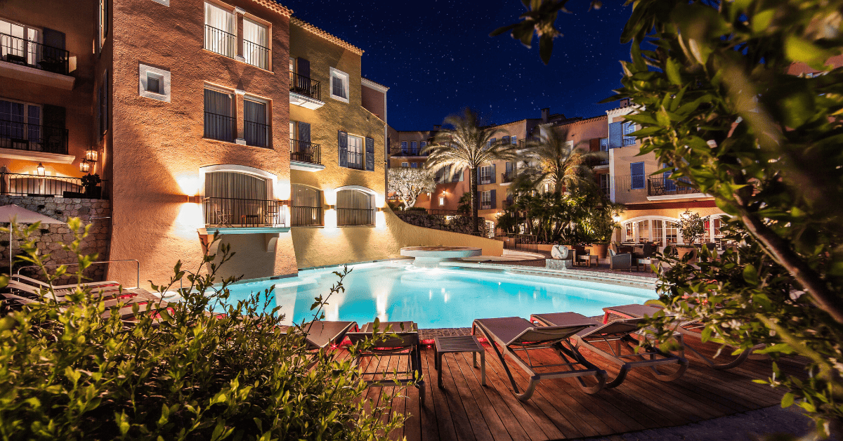 A serene night-time scene at a luxurious hotel, showcasing a beautifully illuminated swimming pool surrounded by comfortable lounging chairs, with the elegant architecture of the hotel and lush greenery enhancing the atmosphere of relaxation and luxury.