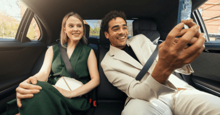 A man and woman seated in the back of a car, smiling while taking a selfie